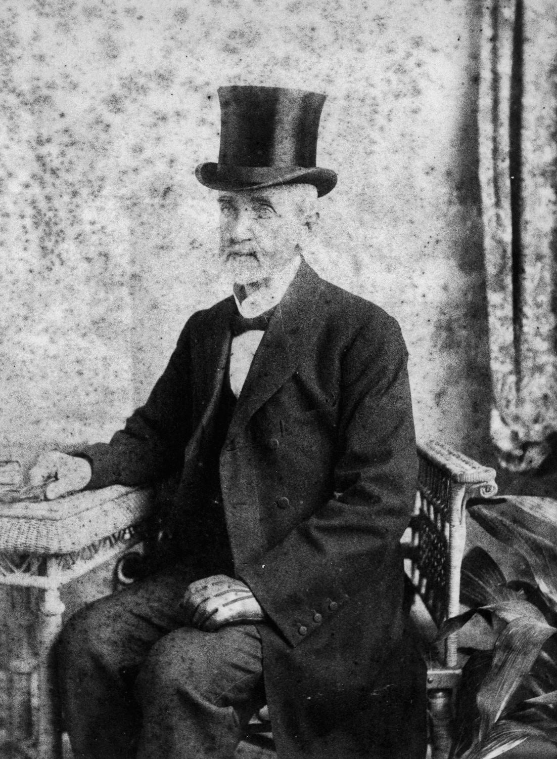 Portrait of man sitting at table wearing top hat and suit with long coat