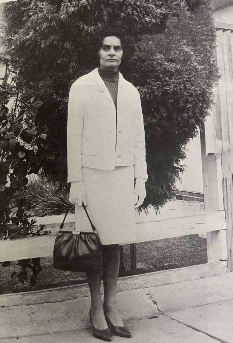 Woman standing in front of a bush wearing a light coloured suit jacket and skirt, gloves and a dark turtle neck top, holding a dark handbag in her right hand, c. 1960s