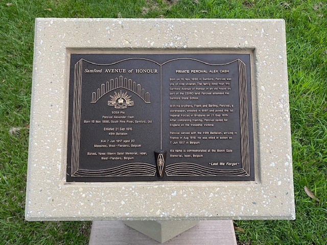 Small black plaque with text designed to look like pages of an open book set in white stone plaque sitting upon grass