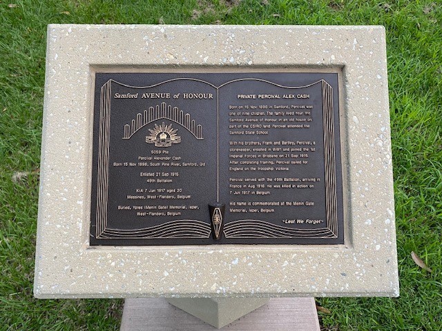 Small black plaque with text, designed to look like pages of an open book, set in white stone plaque, sitting upon grass.