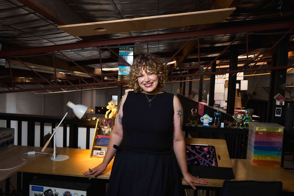 Curly-haired woman with tattoos on arms standing at desk on mezzanine in warehouse studio
