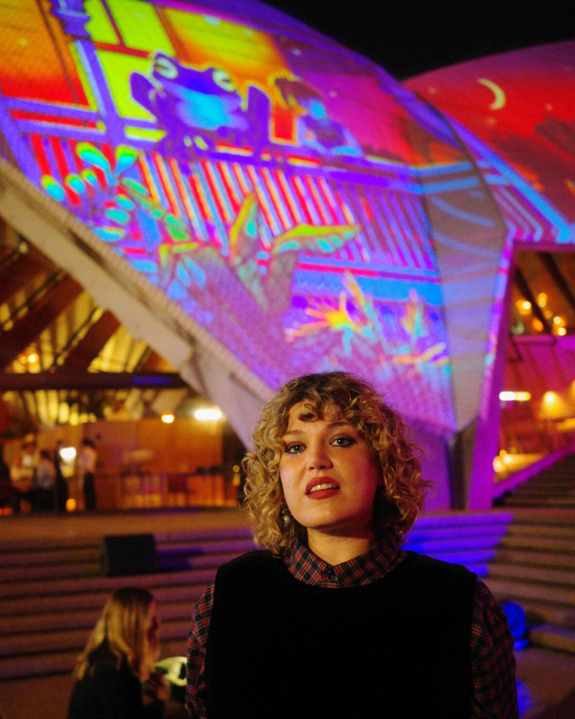 Woman with curly blonde hair standing in front of Sydney Opera House sail illuminated with a projection of a frog and girl on verandah of Queenslander