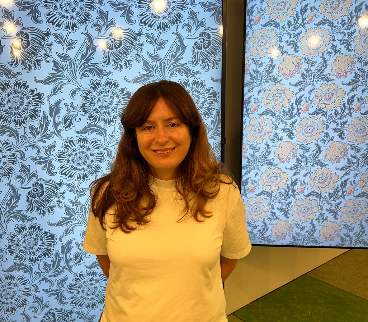 Anna Rowe, in a white t-shirt stands in front of two screens that showcase her floral wallpaper work. Her arms are behind her back and she is smiling at the camera, her hair is around her shoulders.