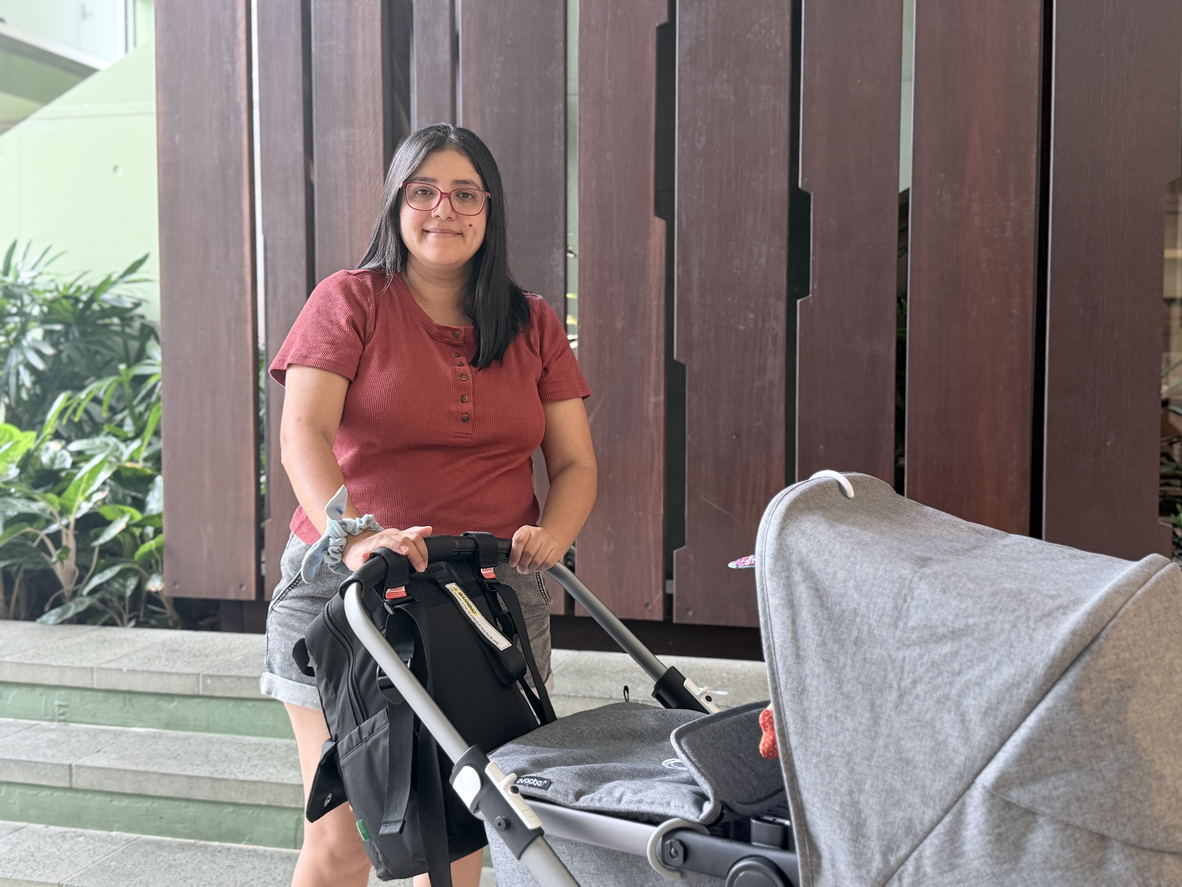 A woman with long black hair and red glasses stands in front of some dark wooden slating holding the handle of a grey pram.