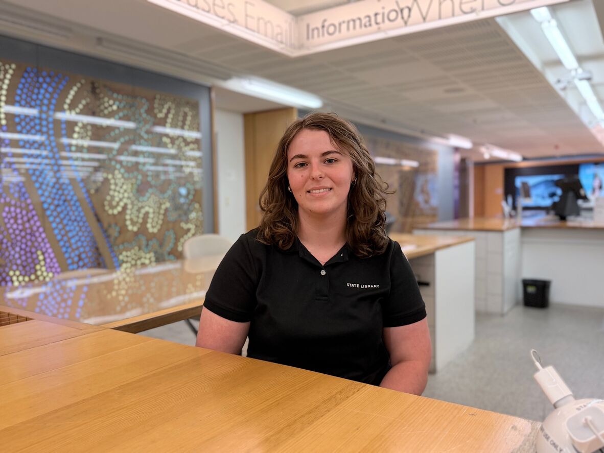 Tara sits at a wooden desk in front of a colourful light background, she is wearing a black state library uniform top and has shoulder length brown curly hair. She is smiling at the camera.