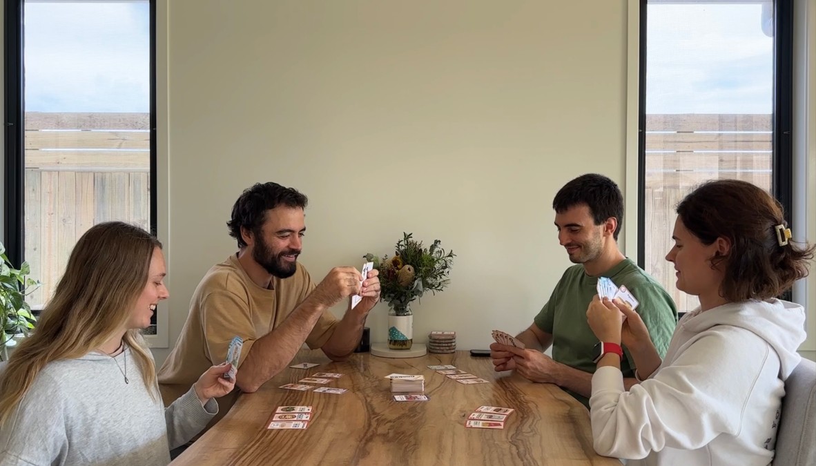 The four Cooked game developers sitting around a light coloured table, in the middle of the table there are a bunch of the Cooked game cards and each person is holding a bunch of cards.  