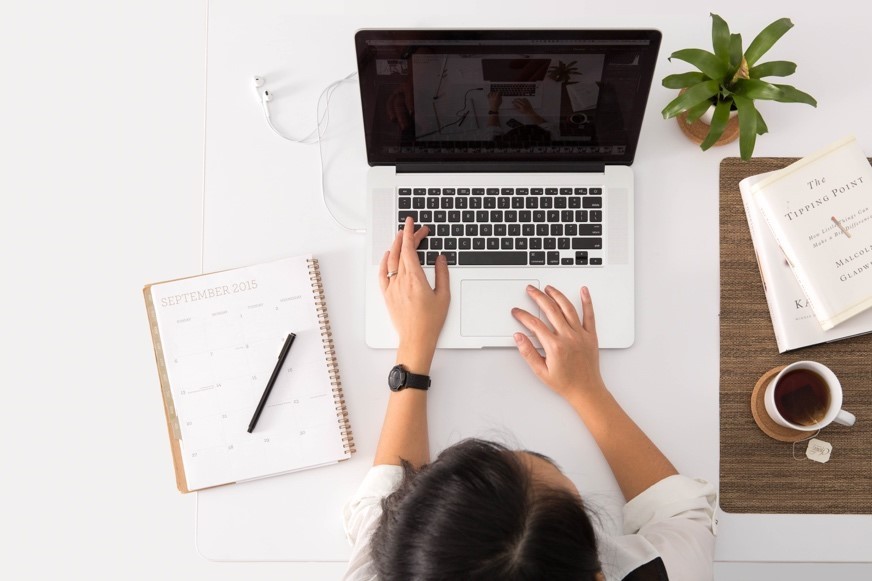 Picture from above of a girl at a laptop with a notebook, pen, plant and cup of tea at her desk