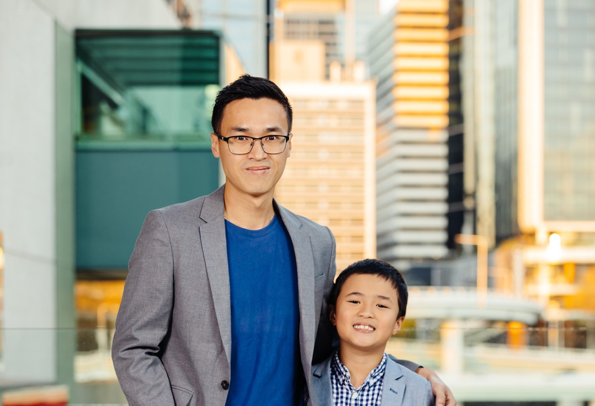 Siang Lu stands in a blue shirt and blazer with his arm around his son Behind them is skyline of Brisbane at sunset them 