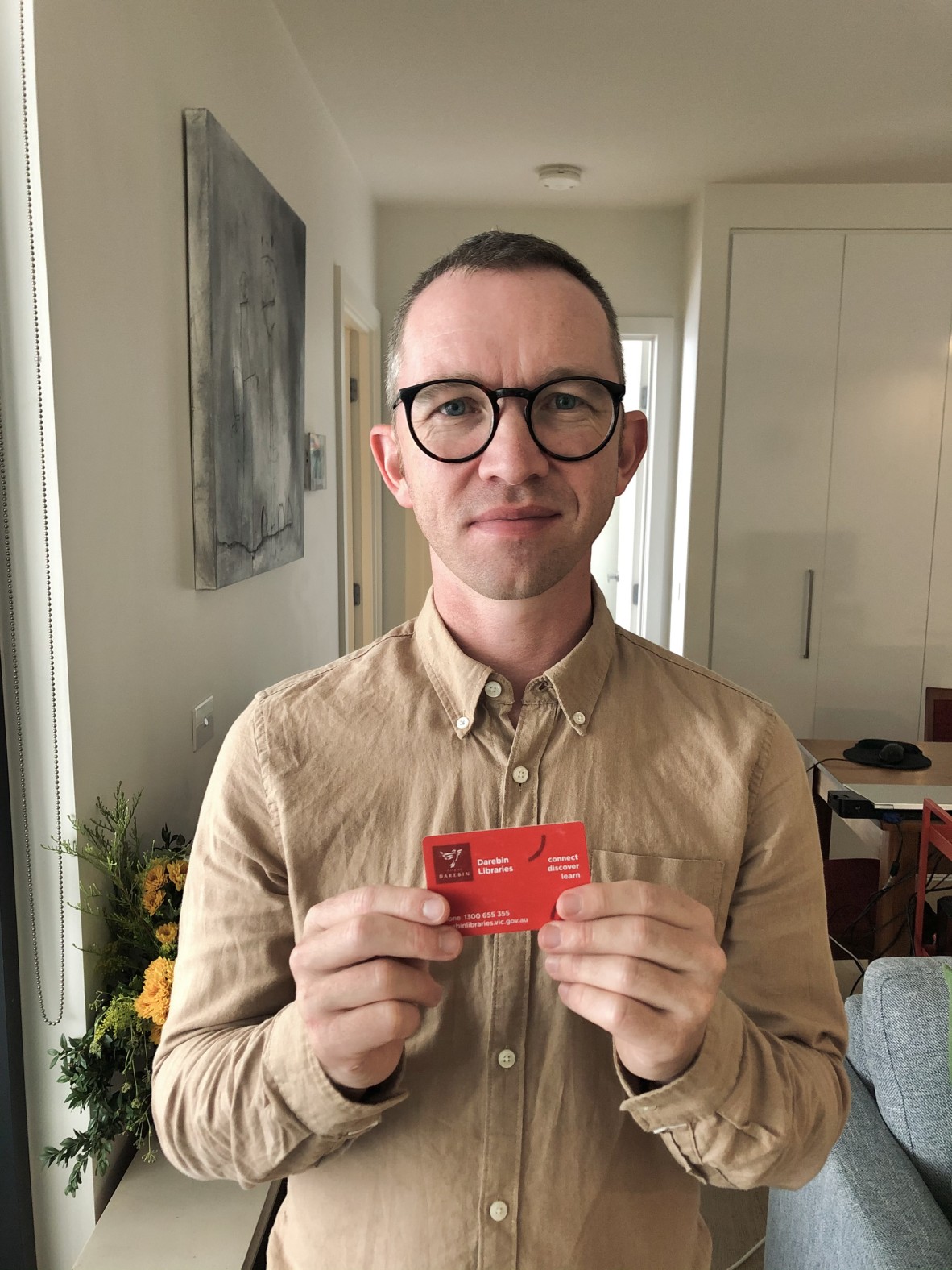 Robert holds up a red library card in the background is a vase of flowers and a couch and table