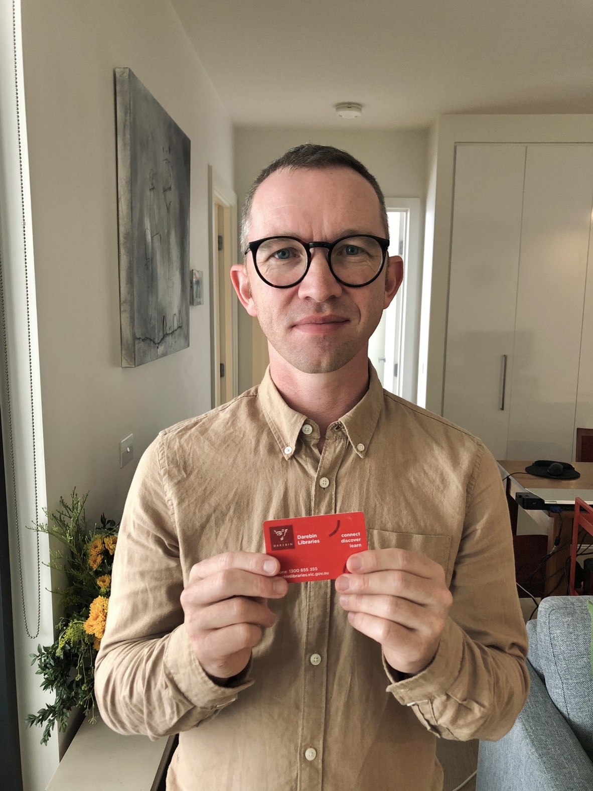 Robert holds up a red library card; in the background is a vase of flowers and a couch and table