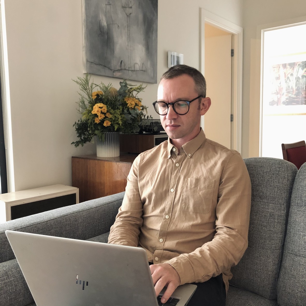 Robert Lukins sits on a couch, typing on his laptop; Robert wears glasses and a brown button down shirt