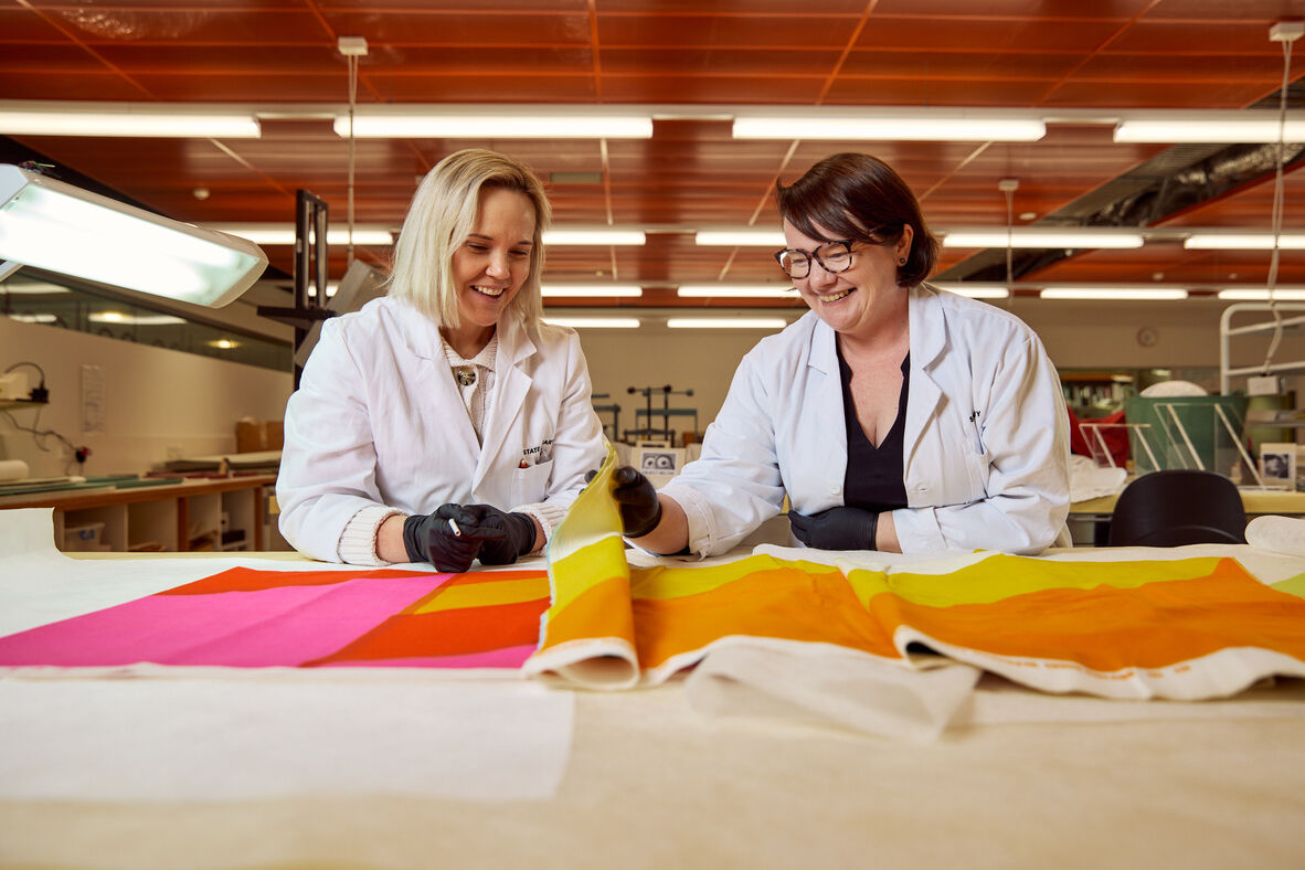 Two women in lab coats and black gloves looking at colourful fabrics on a lab bench