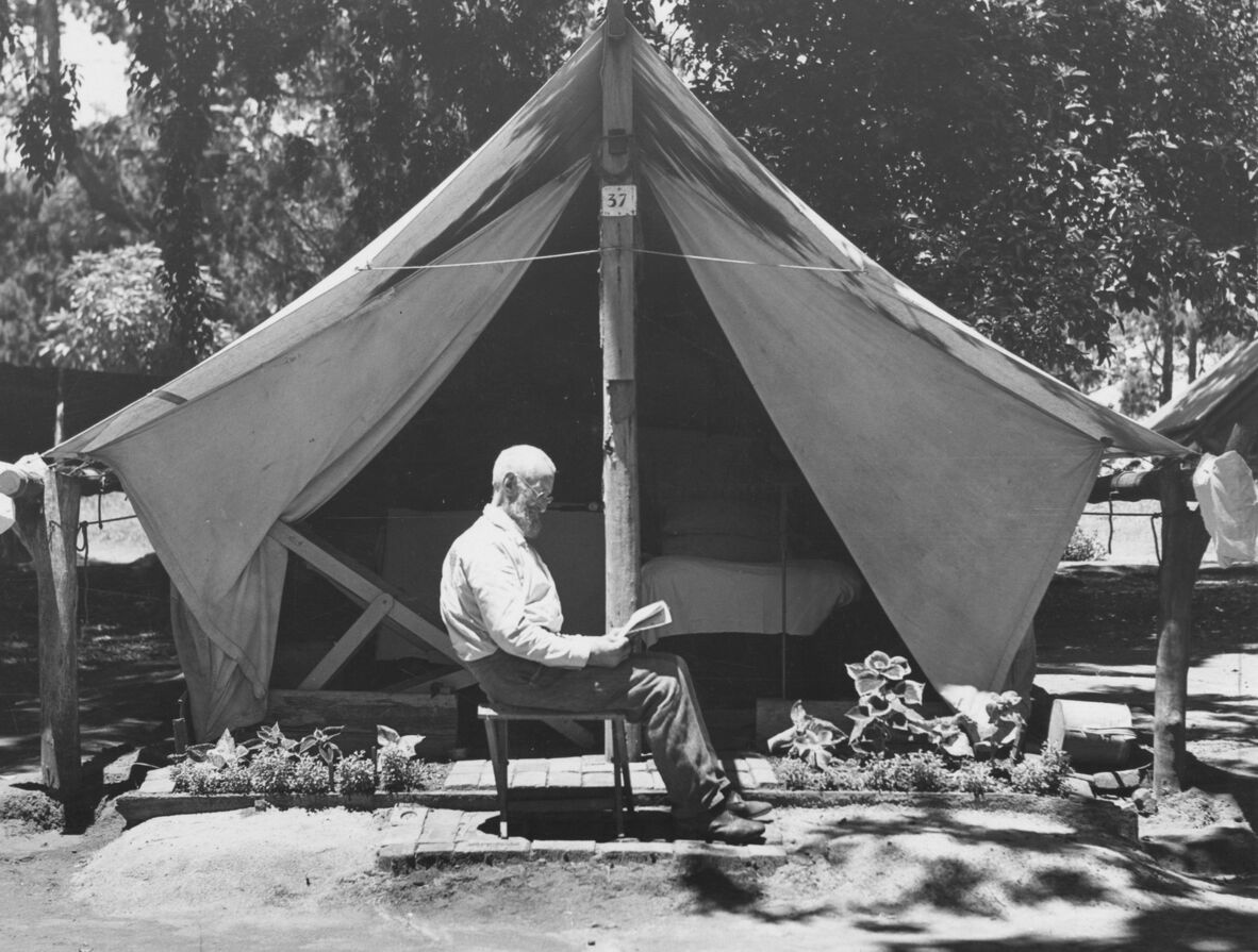 Black and white photo of a man sitting on a stool reading outside a tent