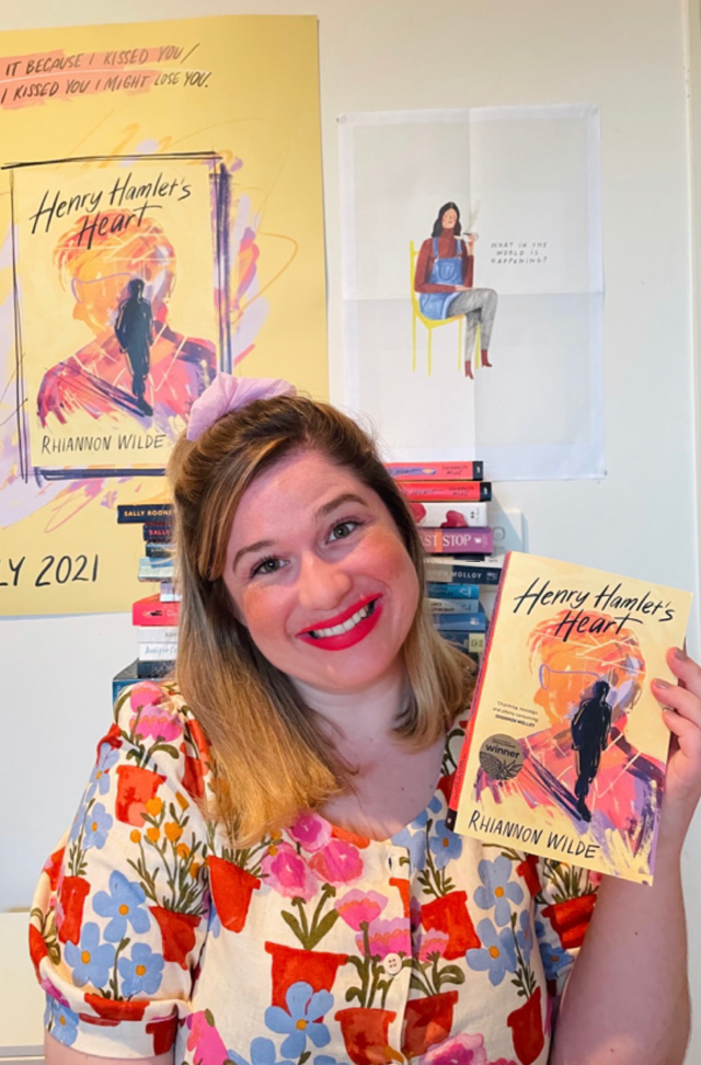 Rhiannon sits in front of a poster of her book cover. She wears a bright floral shirt and holds up a copy of her novel.