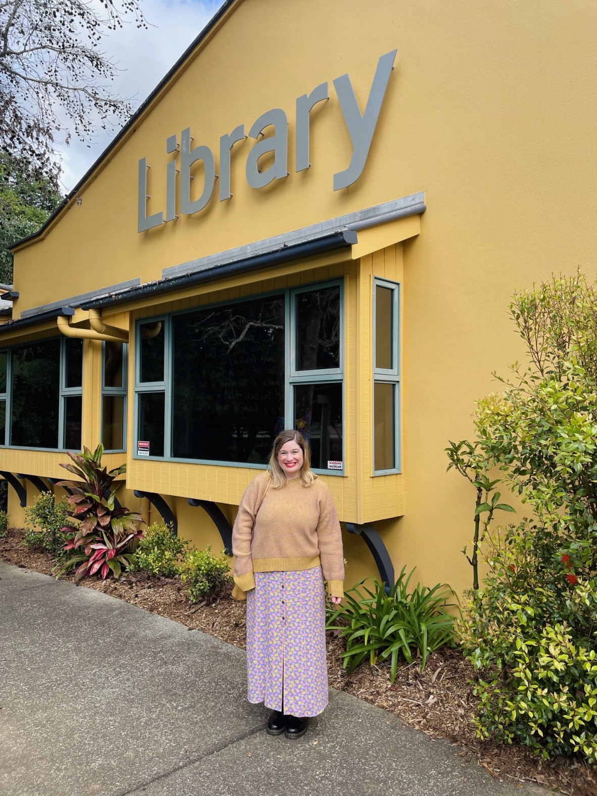 Rhiannon smiles in a yellow jumper and long purple skirt; she stands in front of a yellow building that says 'library'
