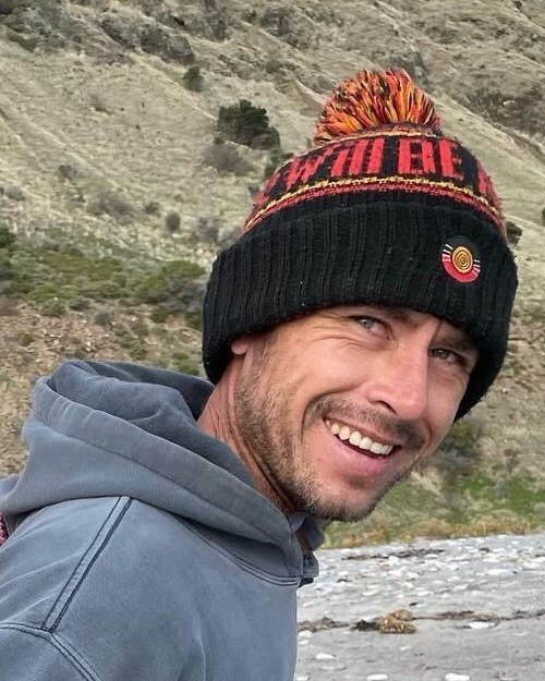 Photo of Rick Slager  he is wearing a grey jumper and black beanie with an Aboriginal flag on it Rick is in front of rocks and grass and is smiling at the camera