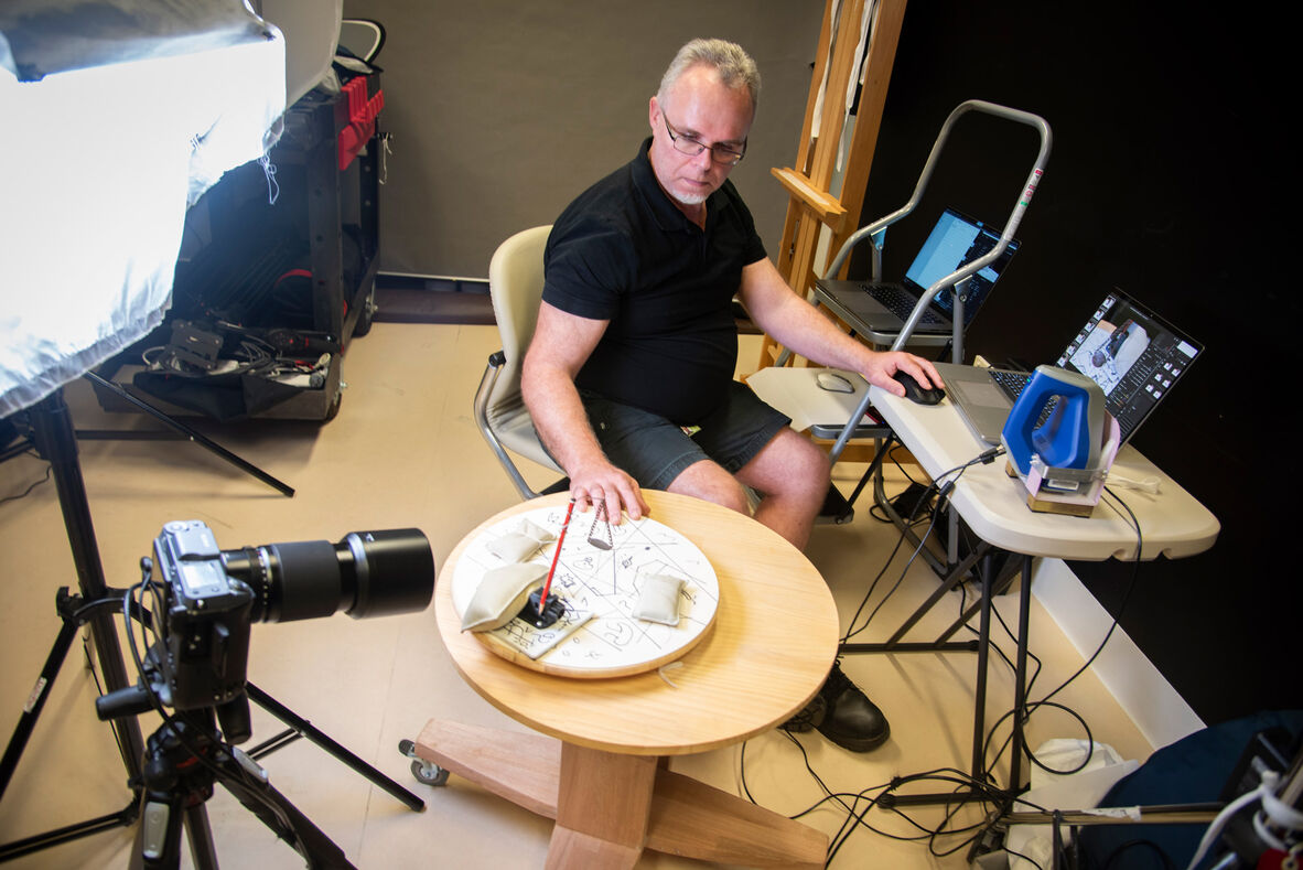 Man sitting on chair with hand on computer mouse while using 3D scanning equipment on old ID bracelet suspended on table