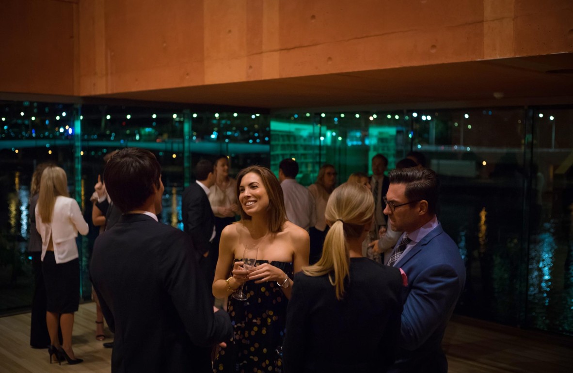 A group of people gathered in the Red Box, with the nighttime river scene visible through the large window.