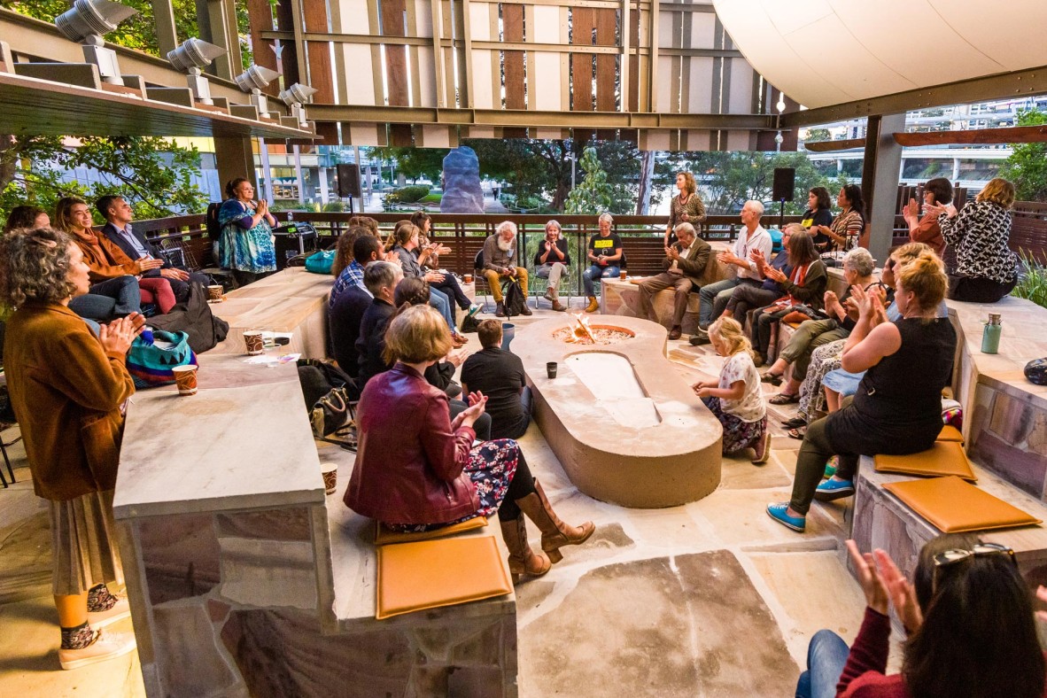 A group of people gathered around a fire pit in an outdoor space, listening to speakers.