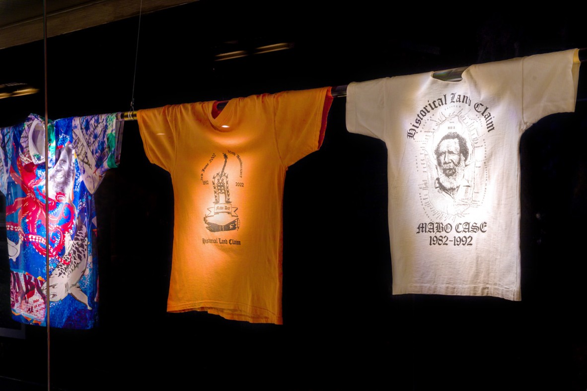 Three shirts with prints celebrating the Mabo High Court case hanging by their sleeves on a pole in a glass case