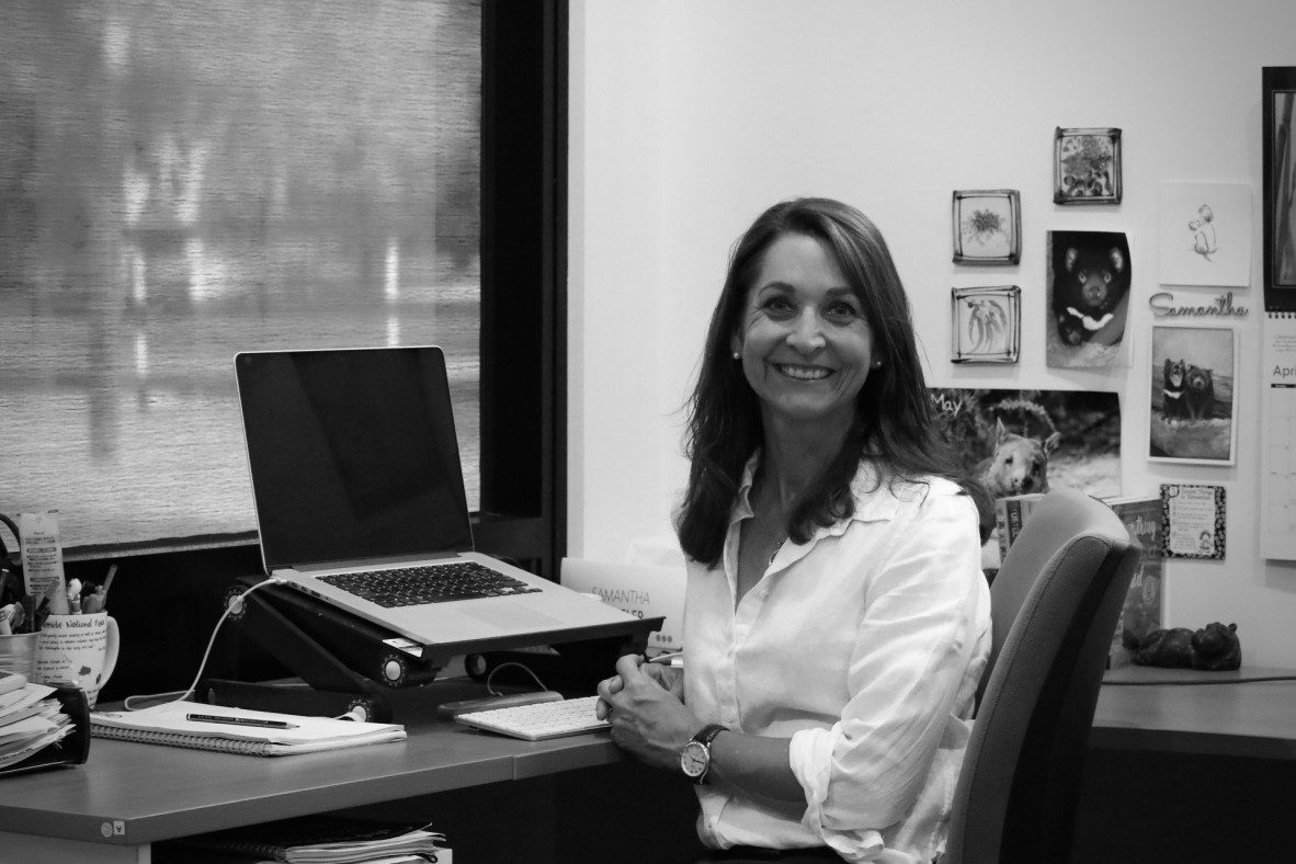Black and white photo of Samantha Wheeler at her office desk She wears a white shirt and is smiling