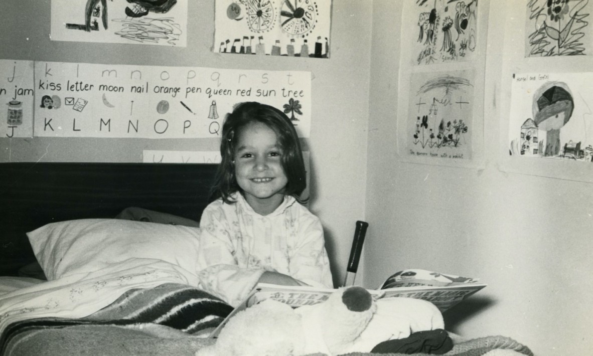 A black and white pic of Samantha Wheeler as a four year old She sits in bed with posters around her a tennis racquet and shes smiling and reading