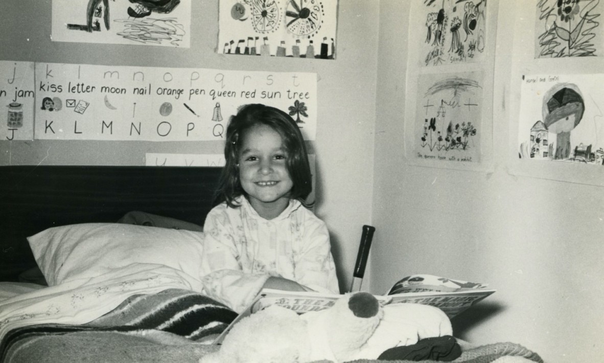 A black and white pic of Samantha Wheeler as a four year old. She sits in bed with posters around her, a tennis racquet and she's smiling and reading