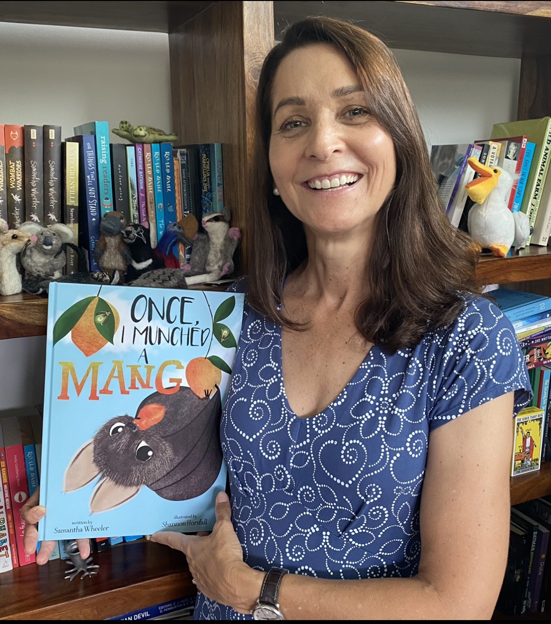 Samantha Wheeler smiles in front of a bookshelf she holds a copy of Once I Munched a Mango