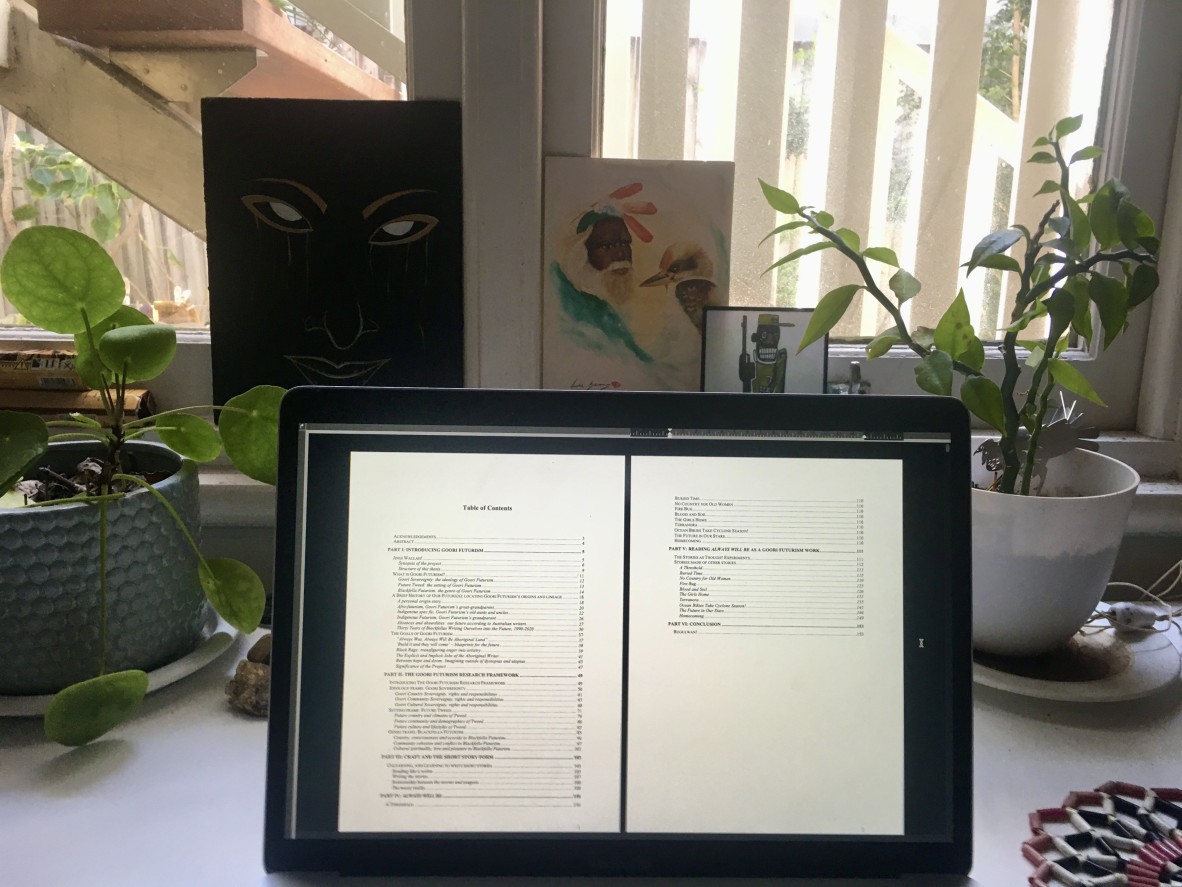 On a desk with pot plants and artwork is an iPad showing the contents page of Mykaela Saunders PhD thesis