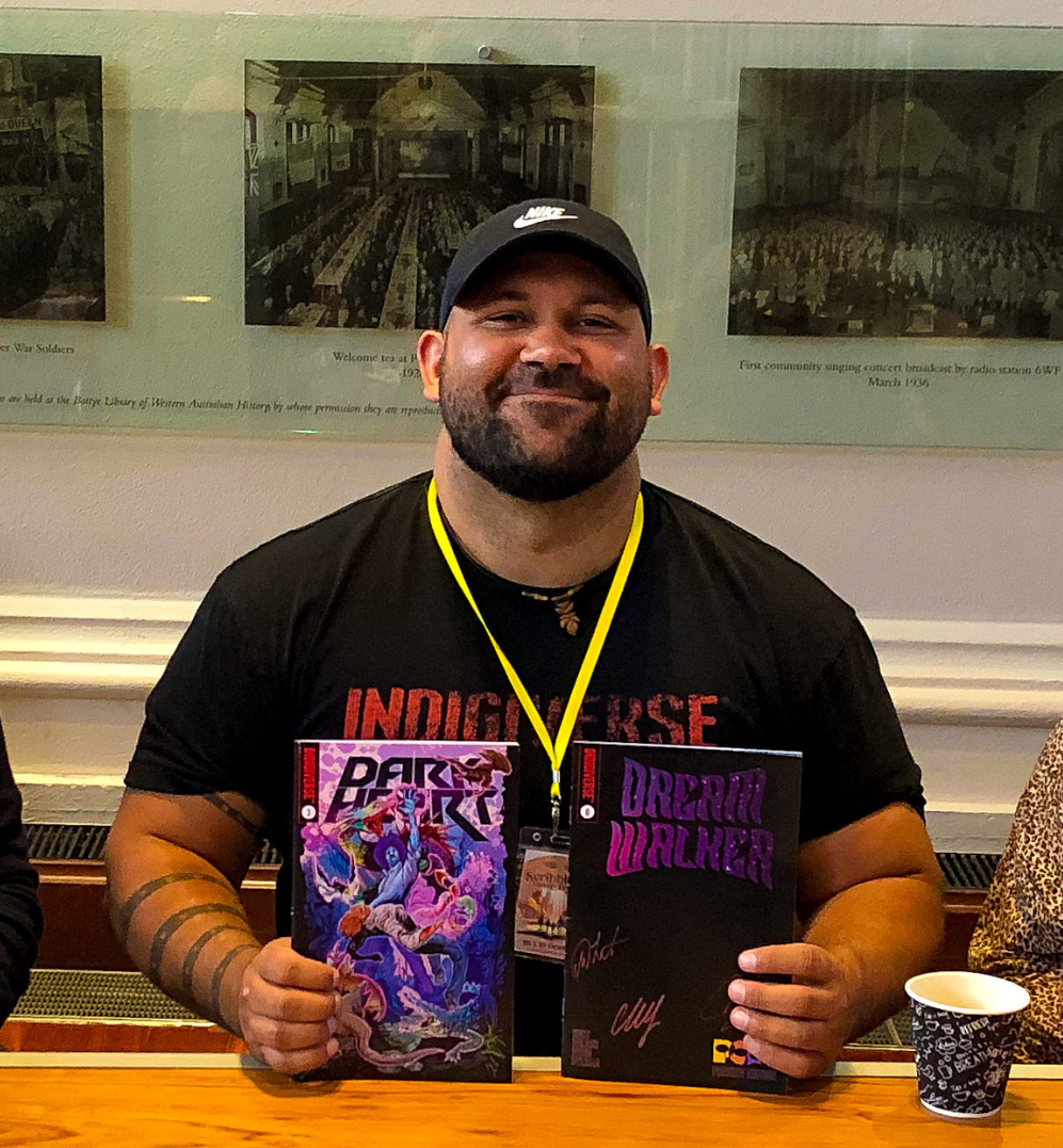 A smiling man with brown skin wearing a black cap and t shirt holding up two comic books . His shirt has INDIGIVERSE written on it.