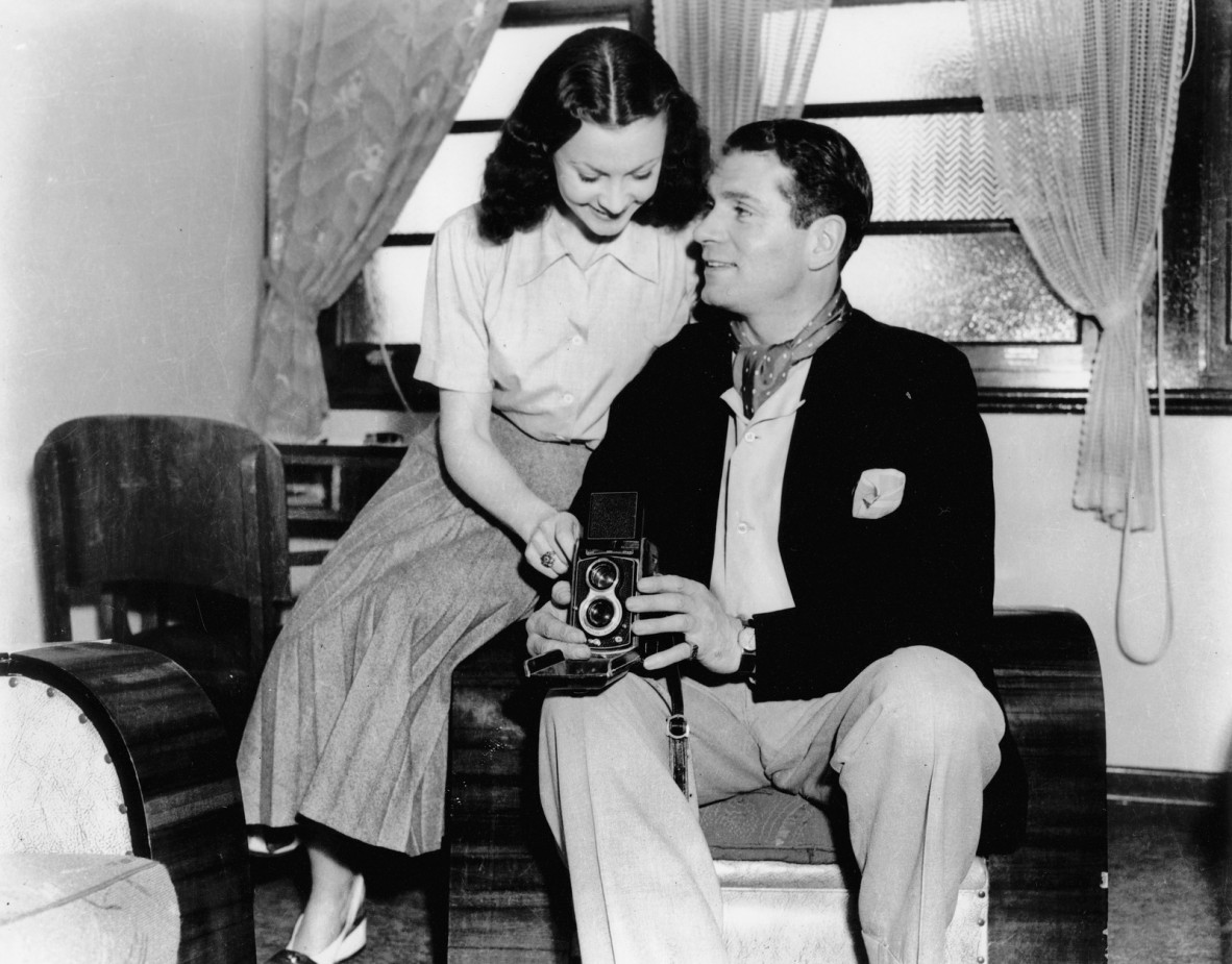 Sir Laurence Olivier and Lady Olivier, (Vivien Leigh) on a short holiday at the Gold Coast after touring with the Old Vic Company in Hobart. Sir Laurence is elegantly dressed in a jacket, flannels and a open-necked shirt with a cravat. Lady Olivier is wearing sunray pleated skirt and a short-sleeved blouse. They are posing with a camera.