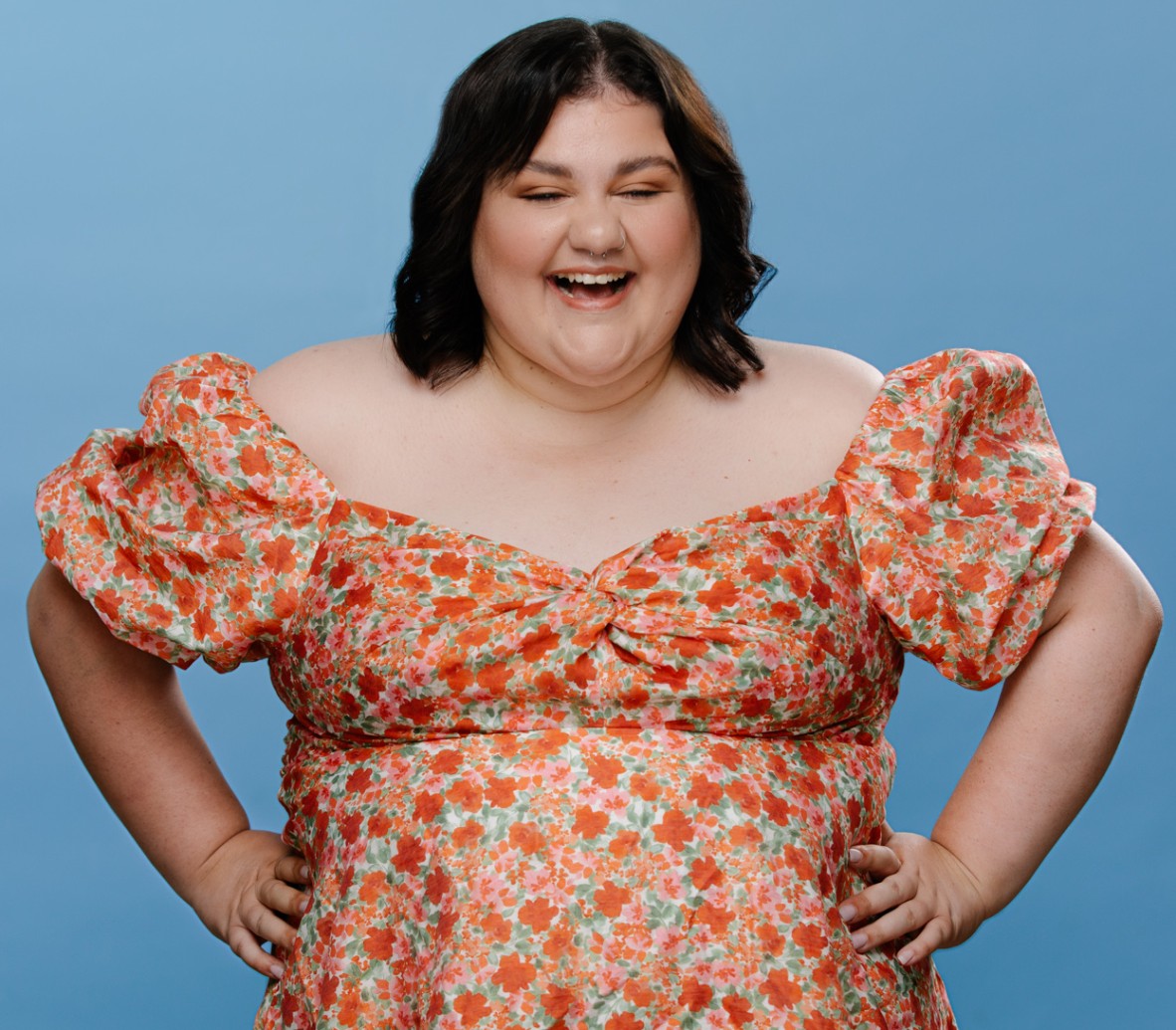 A smiling woman with medium length brown hair wearing a multicoloured dress, standing with hands on hips in front of a blue background.