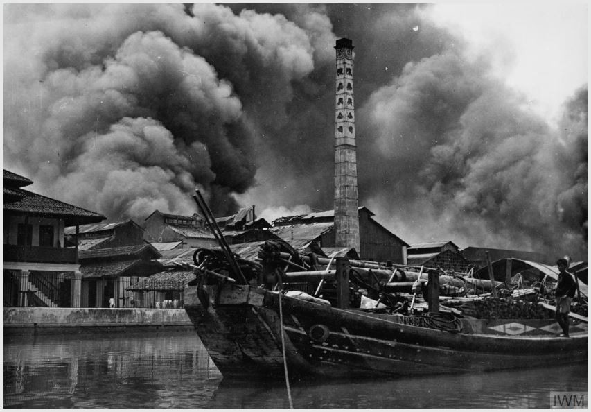 smoke billows from the shore while a old ship is moored on the water 