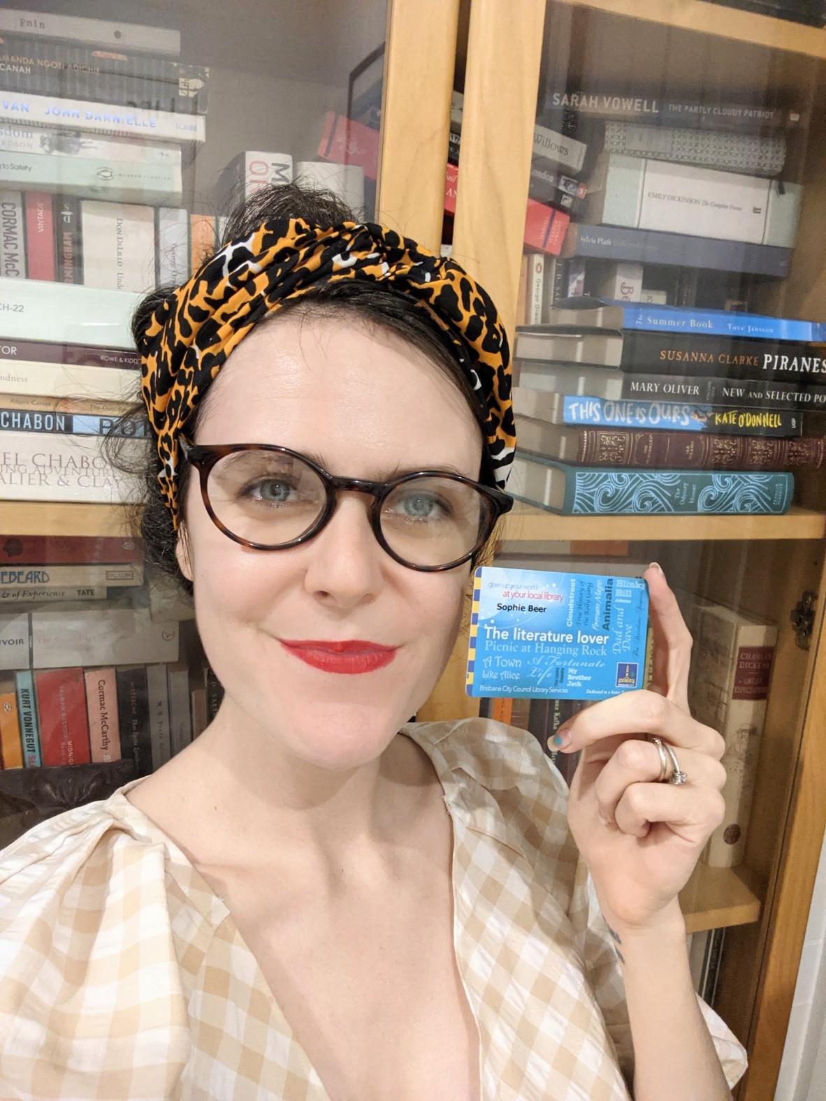 A close up of Sophie Beer who wears a headband and stands in front of a book shelf, holding her library card.