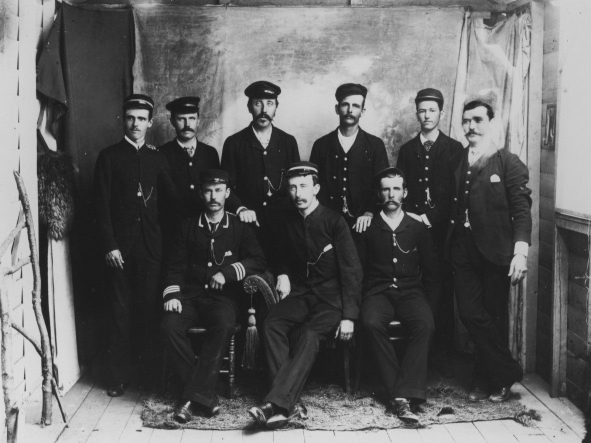 Black and white photograph of 9 uniformed men. Six are standing and three are seated in front.