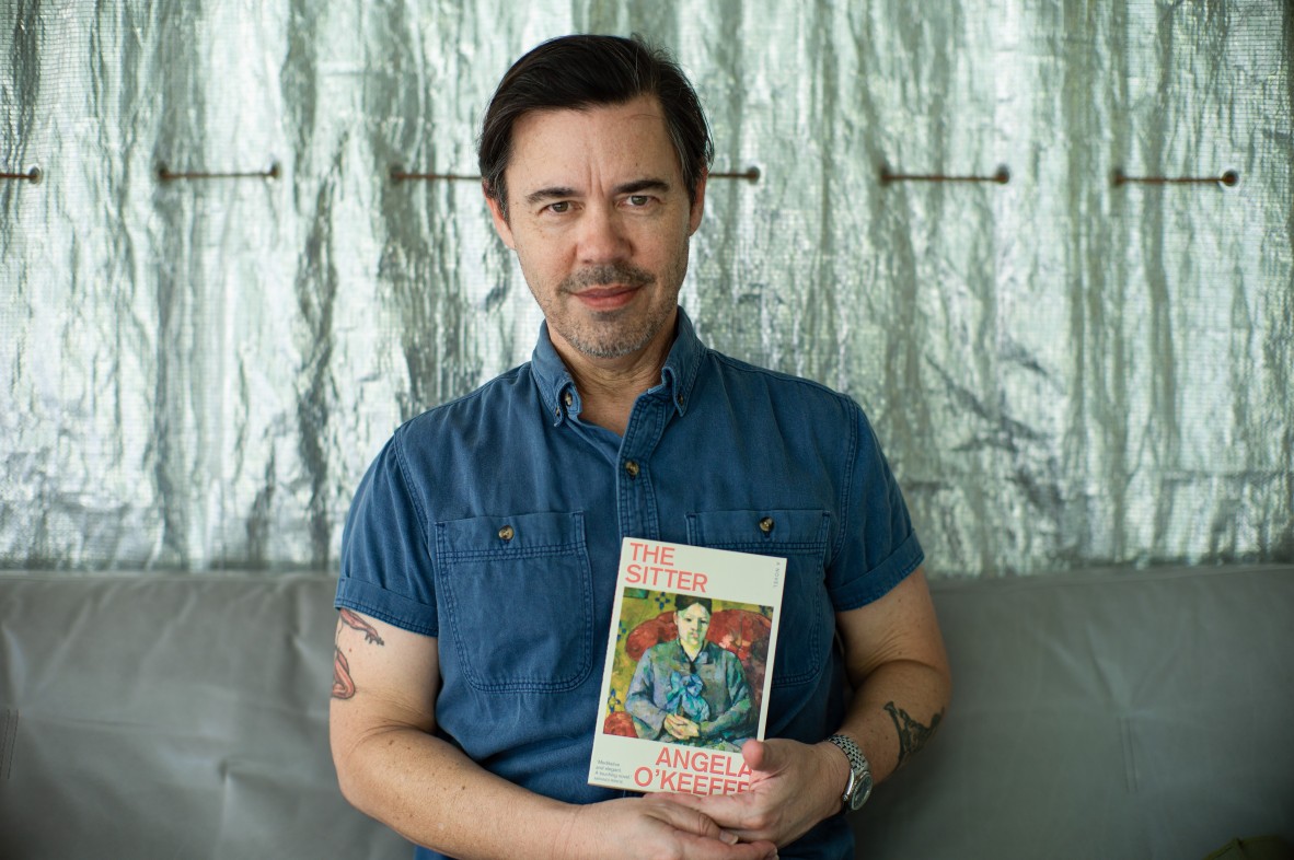 Steve MinOn sits on a couch holding a copy of The Sitter by Angela O'Keeffe