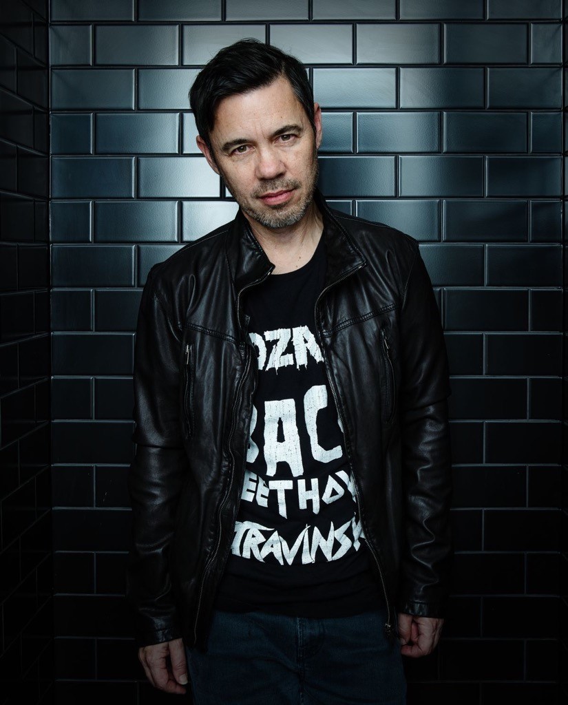 Steve MinOn stands against a wall of dark tiles. Steve wears a black leather jacket.