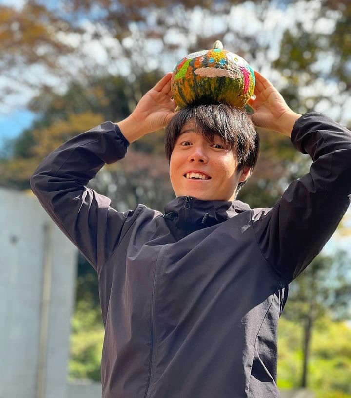 A Japanese person wearing a navy blue jacket smiles and holds a painted pumpkin on their head