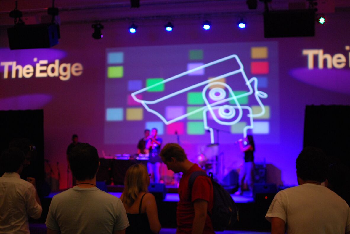A photo of a band playing in the background and crowd in forground at The Edge