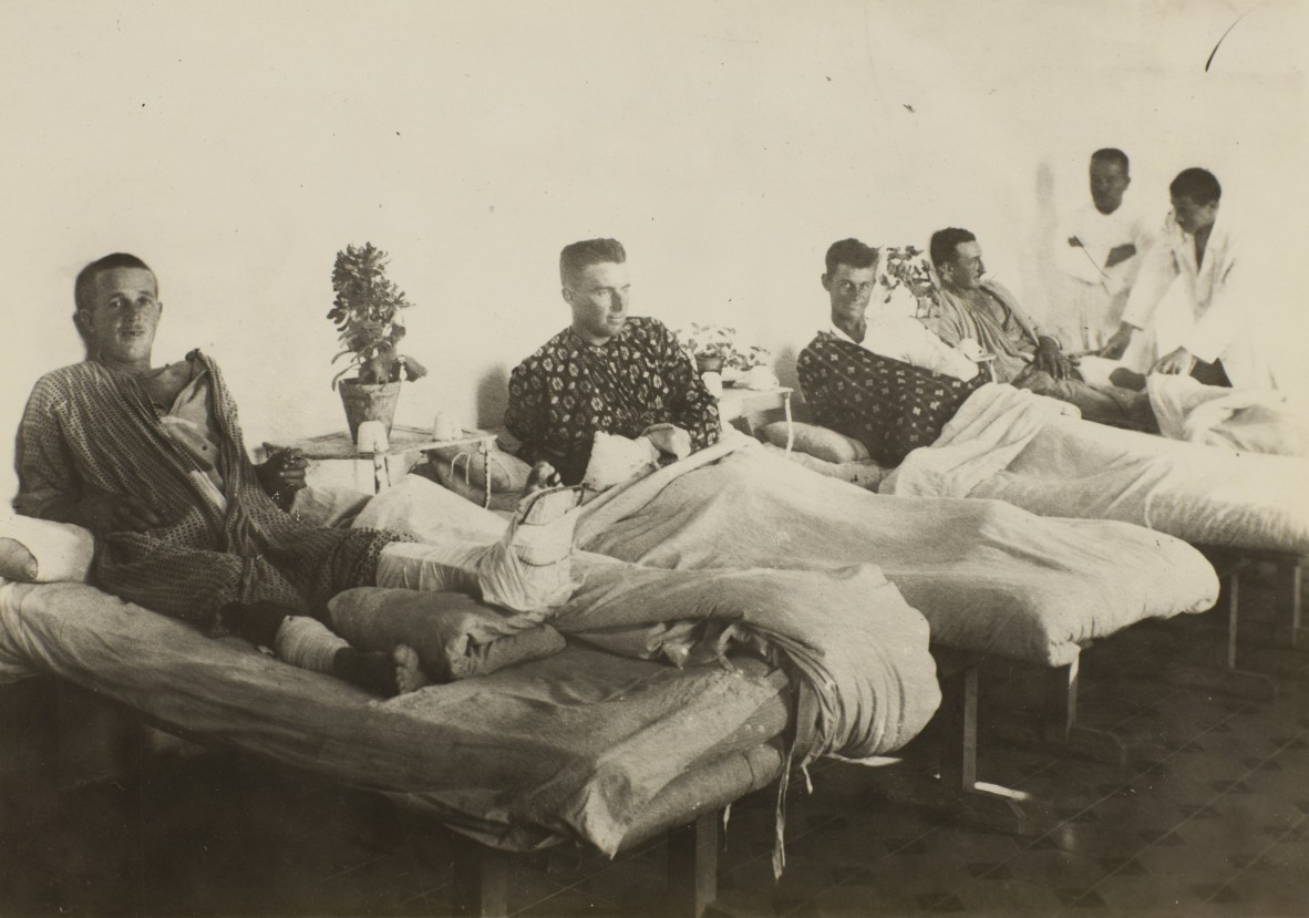 Four men in a hospital ward in beds lined up against a wall being attended by two men in white coats The man in the foreground has his left leg in a splint