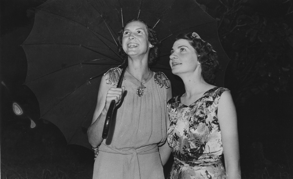 A black and white photograph of two women in dresses standing under an umbrella