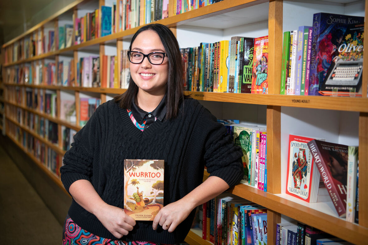 Young woman with dark hair and glasses standing next to bookshelf and holding book with word Wurrtoo on the cover