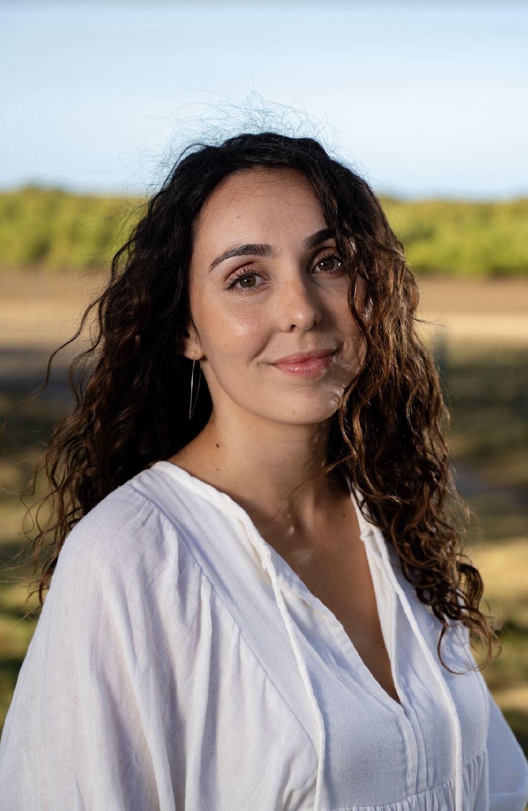 A smiling woman with long dark curly hair in a white top standing with the sky and trees in the far background.