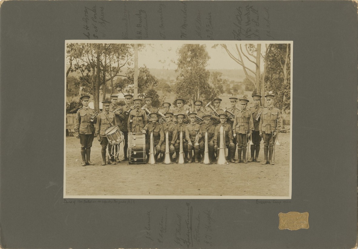Group photo of twenty soldiers in uniform holding brass instruments and drums trees in background dirt ground cover 