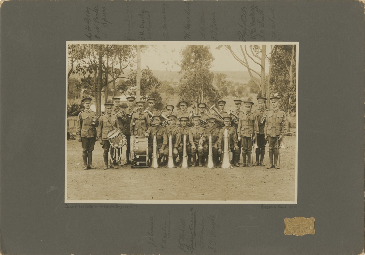 Group photo of twenty soldiers in uniform, holding brass instruments and drums, trees in background, dirt ground cover.