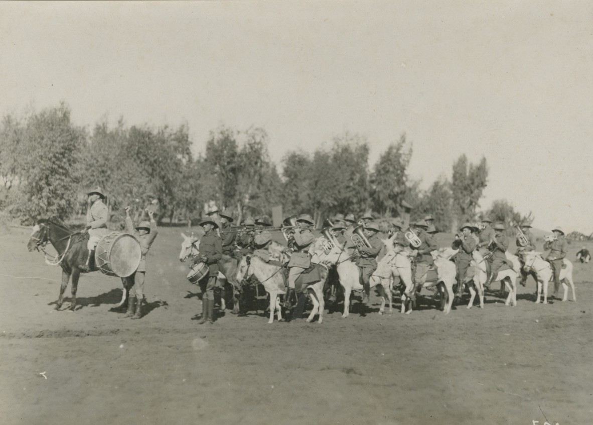 Group of approximately 20 soldiers in uniform, facing left of camera point of view, riding donkeys. Soldiers playing brass instruments and drums.