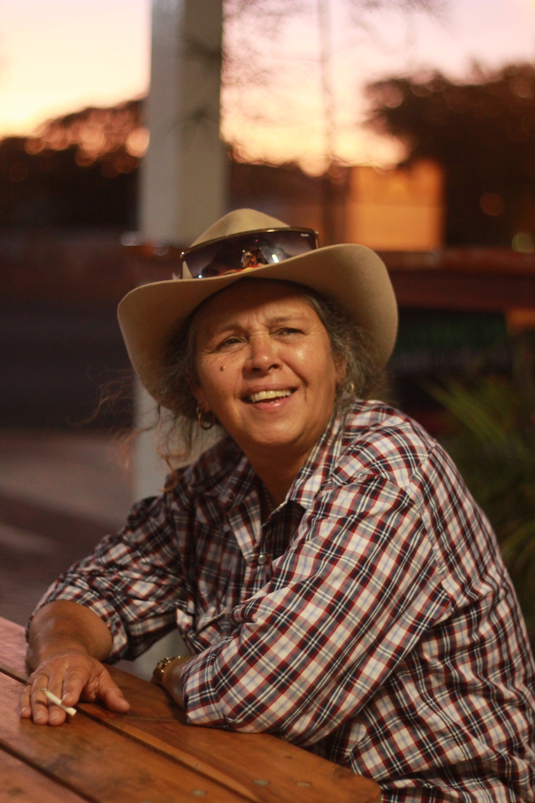 Keelen Mailman OAM sitting at a table at the pub, wearing her hat with a cigarette in hand 