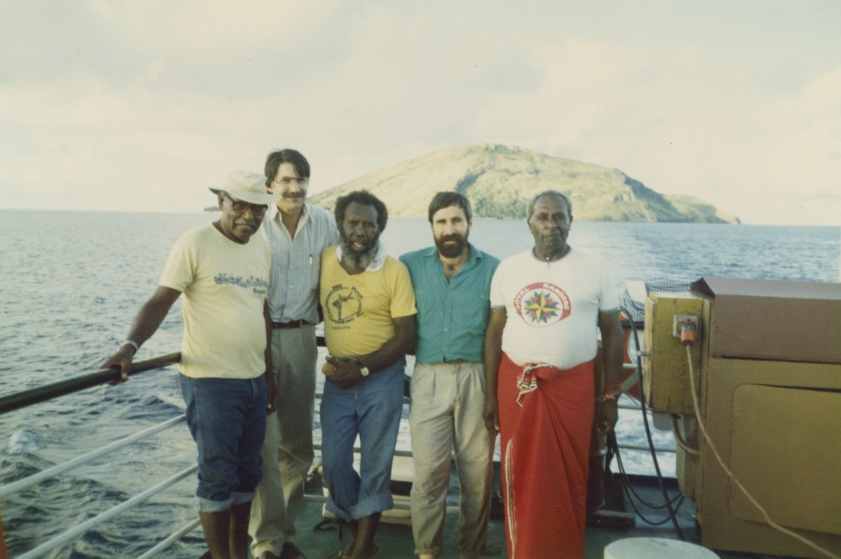 5 men stand on a boat in front of an island 
