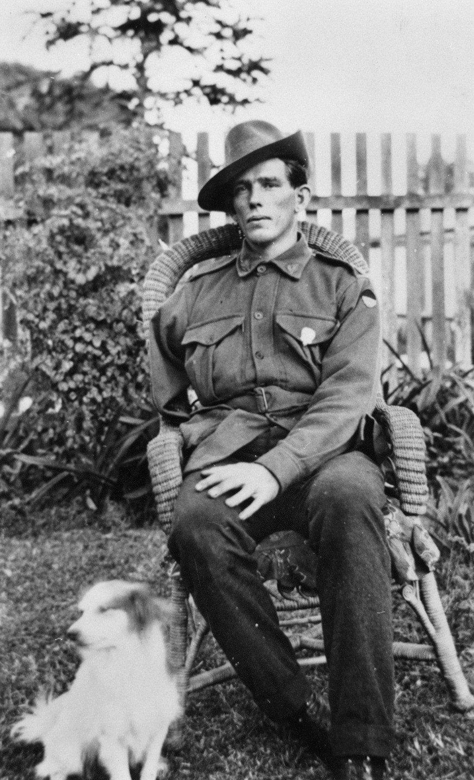 Man in uniform, appears to be missing all or part of right arm, sitting in outdoors in cane chair. Dog sitting on ground by his right side.