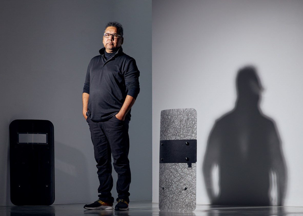 Artist Vernon Ah Kee in dark clothing standing in a grey room with two of his riot shield art works.